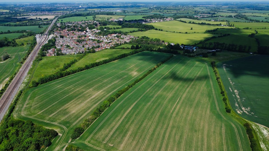 An aerial view of a rural landscape featuring large, well-maintained green fields with visible mowing lines, bordered by neat hedgerows and mature trees. In the background, a small residential area wi